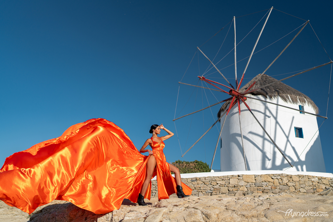 A woman in a flowing orange dress poses on a rocky hill in front of multiple traditional windmills in Mykonos, with the wind catching her dress and a clear blue sky overhead.